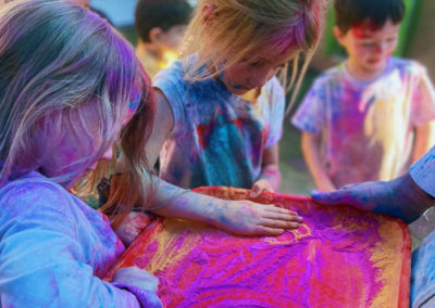 Two blond-haired girls rub their hands through a red tray filled with brigh fuschia powder while a little boy stands looking down at something in the background.