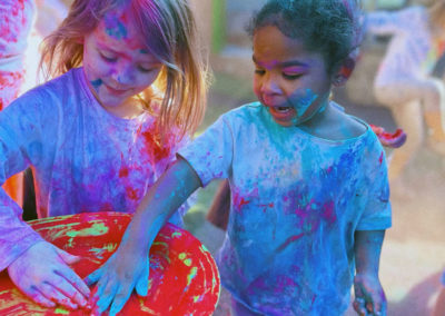 Two little girls, one Caucasian and the other African American, stand side by side rubbing their hands in a tray filled with brightly colored powder. They are covered in bright powder as well.