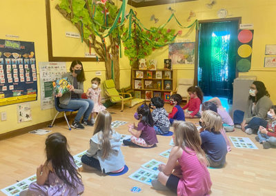 Storytime at Lanugage Garden Preschool with children sitting cross-legged on the ground facing a teacher holding up a book with a little girl next to her. The room is painted a bright yellow with paper crafted trees on the walls.