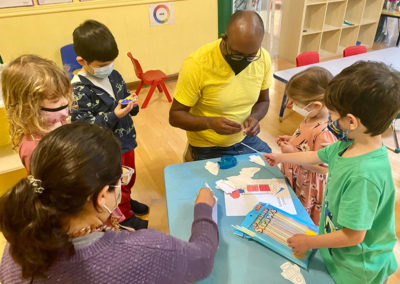 Classroom at Language Garden Preschool with bright yellow painted walls. The teacher wears a bright yellow shirt and kneels around a low blue-colored table with children crafting with paper and straws.